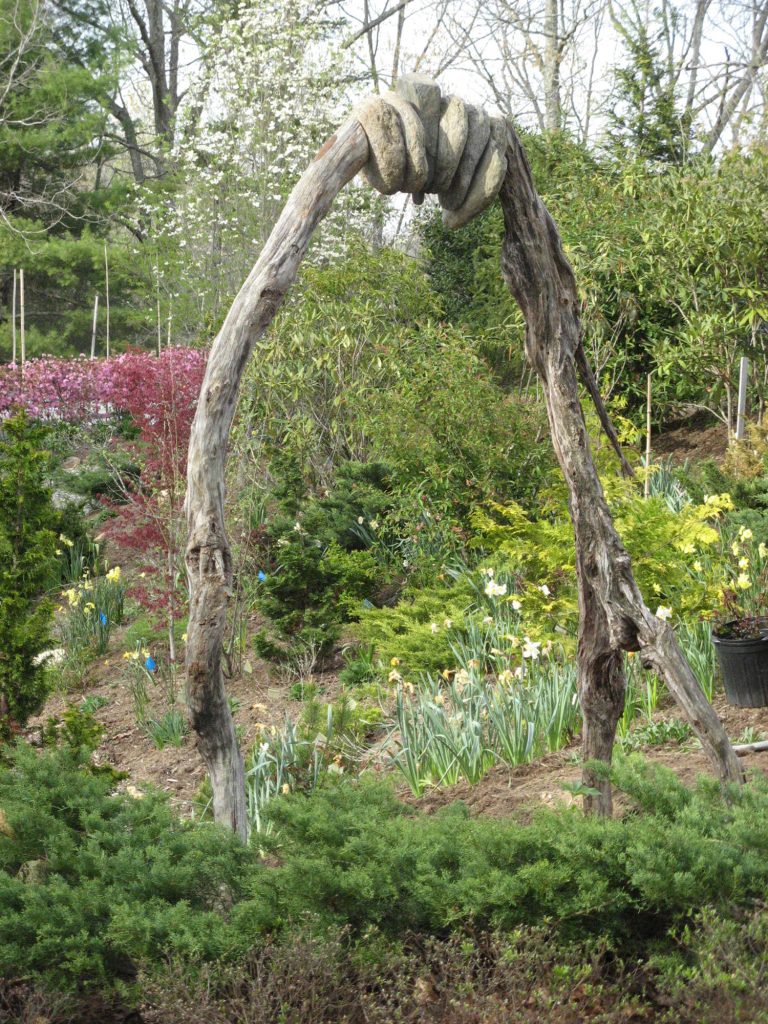 Rock Bridge Prototype by Robert Levin. Two large branches stand vertically bracing six large stones. This natural sculpture stands among a garden in Burnsville, NC.
