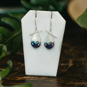 Summer Day in the Blue Ridge Mountains, Ilene KayA pair of earrings hang on a white display. The earrings are a matching set, metal drops with details of blue mountains and yellow stars.